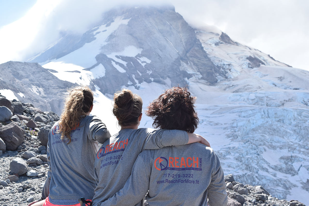 Trio of women wearing Reach Fitness hoodies sitting atop a mountain staring out over Mt. Hood.